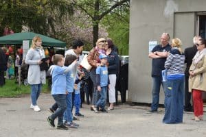 Grupa osób stoi przed budynkiem. Wśród nich są również dzieci. Chłopiec po lewej stronie ma wyciągniętą rękę oraz palec i pokazuje na coś. Za osobami po lewej stronie stoi mały namiot.