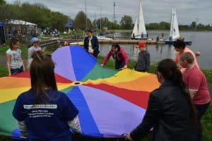 Grupa osób stoi w kole. Są to dorośli i dzieci. Trzymają oni tęczowe koło z materiału.