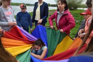 Osoby stoją na zewnątrz koła i trzymają tęczowy materiał. W środku siedzi skulona dziewczynka. Kobieta po prawej stronie ma otwarte usta.