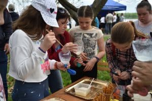 Dzieci stoją przy stole, na którym są tacki, paluszki. Niektóre dzieci trzymają ucięte butelki a na otworze są założone balony. Kobieta po lewej stronie stoi obok dziewczynki i trzyma taką butelkę.