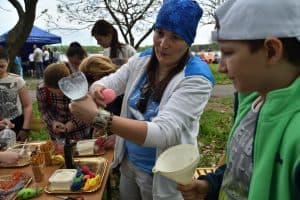 Chłopiec stoi po prawej stronie i trzyma w ręku lejek. Obok niego stoi kobieta, która w wyciągniętej ręce trzyma uciętą butelkę i gąbkę. Na stole leżą paluszki, tacki z balonami.
