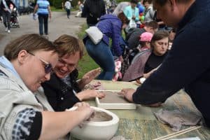 Przy drewnianym stole siedzą dorosłe osoby. Kobieta po lewej stronie ma przed sobą miskę z gliną, którą dotyka ręką. Obok jest kobieta zwrócona twarzą do niej i uśmiecha się. Po prawej stronie stoi mężczyzna nachylający się do stołu.