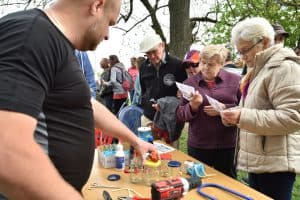 Przy stole stoją ludzie. Po prawej stronie są dwie starsze kobiety, które trzymają w rękach kartki. Po lewej stronie męzczyzna ma wyciągnięta reke i dotyka słoika stojącego na stole. Na stole leżą słoiki, klej, wiertarka.