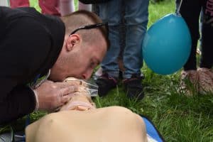 Mężczyzna jest pochylony nad manekinem do ćwiczeń. Obejmuje on swoimi ustami usta manekina a ręką podtrzymuje podbródek. Po prawej stronie są nogi dzieci ucięte przez obiektyw oraz niebieski balon na patyku.