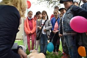 Dzieci stoją na trawniku i trzymają balony. Wszystkie patrzą na kobietę siedząca po lewej stronie. Przed nimi leży fantom.