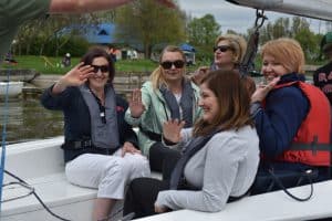 Pięć kobiet siedzi na żaglówce. Każda ma na sobie kapok. Za nimi jest woda, łąka i dom.