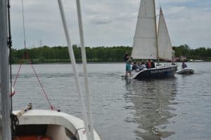 Na wodzie jest żaglówka na której są ludzie. Przed nimi płynie ponton. Po drugiej stronie jest las.