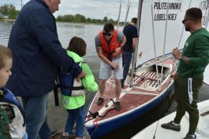 Mężczyzna stoi na żaglówce w kapoku i trzyma się liny. Naprzeciwko niego stoi dziecko, a za nim starsza osoba, która trzyma dziecko. Po prawej stronie stoi mężczyzna bokiem do obiektywu.