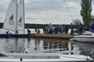 Ludzie stoją na pomoście. Po lewej stronie jest żaglówka na której jest dwoje ludzi. Po prawej stronie również są zacumowane żaglówki.