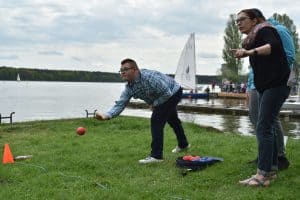 Na trawniku są trzy osoby. Mężczyzna stoi przed linią i jest zgięty w pół. Jedną rękę ma wyciągniętą przed siebie a przed nią leci kula. Po prawej stronie kobieta pokazuje coś palcem i zasłania mężczyznę, który stoi za nią.