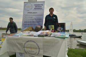 Na trawniku stoi stół, na którym leżą książki. Za stołem stoi kobueta. Obok niej jest baner z napisem: “Pedagogiczna Biblioteka Wojewódzka im. Komisji Edukacji NArodowej. Oferujemy: współpracę z placówkami oświaty, zajęcia edukacyjne, warsztaty metodyczne dla nauczycieli, specjalistyczny księgozbiór o profilu pedagogicznym i psychologicznym, szeroki wybór mediów i multimediów”