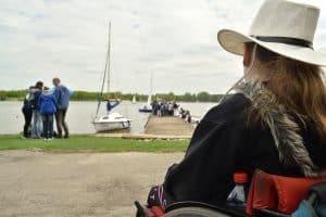 Po prawej stronie siedzi kobieta w kapeluszu na wózku inwalidzkim tyłem do obiektywu. Przed nią jest pomost i żaglówka. Po lewej stronie stoi grupa osób.