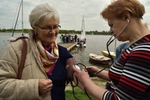 Dwie kobiety stoją naprzeciwko siebie. Kobieta po prawej stronie ma stetoskop w uszach i trzyma pompkę. Kobieta po lewej stronie ma na ramieniu mankiet do mierzenia ciśnienia. Za nimi jest woda i pomost.