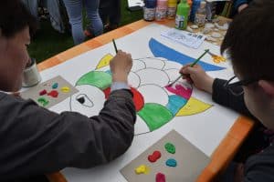 Na stole leży białą kartka z narysowaną rybką. Dwie osoby siedzą i trzymają pędzle. Po części rybka ma już kolory. Osoby sa bokiem do obiektywu i lekko przycięte.