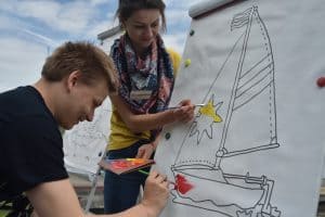 Dwie osoby stoją przy tablicy z namalowaną żaglówką. Jest to chłopak i dziewczyna. Dziewczyna trzyma w jednej ręce pędzel a w drugiej farby wylane na kartonie. Chłopak ma w jednej ręce pędzel.