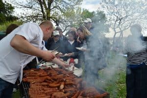 Na dużym grillu smażą się kiełbasy. Po lewej stronie jest kolejka osób. Mężczyzna stoi ze szczypcami za grille. Po prawej stronie stoi osoba przysłonięta przez dym z grilla.