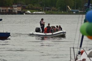 Na wodzie jest ponton z ludźmi. Wszyscy mają kapoki. Po lewej stronie jest żaglówka na której są balony.