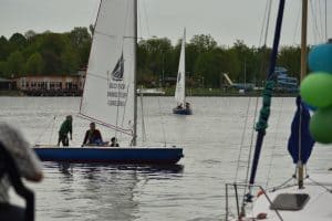 Na wodzie są żaglówki. Po lewej stronie w żaglówce są trzy osoby. Po drugiej stronie są budynki i las.