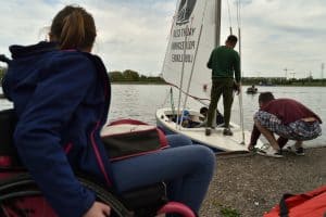 Po lewej stronie jest kobieta na wózku inwalidzkim, która jest tyłem do obiektywu. Po prawej stronie stoi mała żaglówka. Jeden mężczyzna stoi na niej, a drugi kuca.