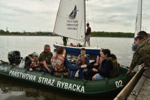 Na dużym pontonie siedzi spora grupa osób. Na pontonie jest napis:” Państwowa Straż Rybacka 02”. Za nią jest żaglówka . Po lewej stronie mężczyzna w moro trzyma rękę na linie.
