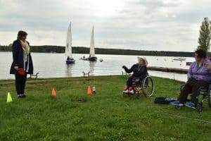 Po lewej stronie na trawniku są dwie osoby na wózkach inwalidzkich. Kobieta pośrodku ma jedną rękę podniesioną do góry. Po prawej stronie stoją pachołki oraz koieta z paletką.