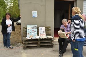 Grupa osób jest przed budynkiem. KObieta po prawej stronie trzyma mikrofon i jest bokiem do obiektywu. Mężczyzna po lewej stronie siedzi i na kolanach trzyma otwartą skrzynkę. Po lewej stronie kobieta opiera się ręką o budynek a w drugiej trzyma telefon. Na wprost na skrzynkach postawiono plakaty.