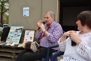 Po prawej stronie siedzą kobieta i mężczyzna. Mężczyzna ma w ręku mikrofon a drugą rękę ma podniesioną na wysokość twarzy. Kobieta patrzy w jego stronę i ma złączone ręce. Po lewej stronie na ławce stoją plakaty.