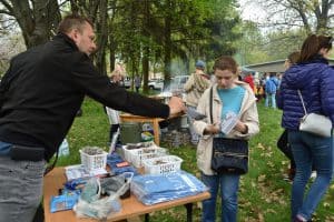 Kobieta stoi po prawej stronie i trzyma broszury w ręce. Po lewej stronie za stołem jest mężczyzna, który ma wyciągniętą rękę z broszurą w stronę kobiety. Na stole leżą koszulki, skrzynki.