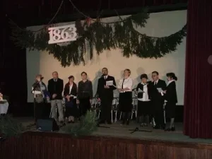 Ludzie stoją na scenie. U góry wisi 1863. Przy ustach maja mikrofony,