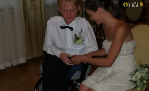 Kobieta w białej sukience trzyma za dłoń mężczyznę na wózku w białej koszuli. Ich twarze są zwrócone ku ich dłoniom.