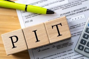 Kostki na których napisane jest “PIT”. W tle papierowe zeznania o wysokości dochodów i papiery do wypełniania PIT-u.