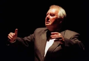 Mężczyzna stoi. Ma podniesione ku górze dłonie. Jego twarz skierowana jest na wprost.