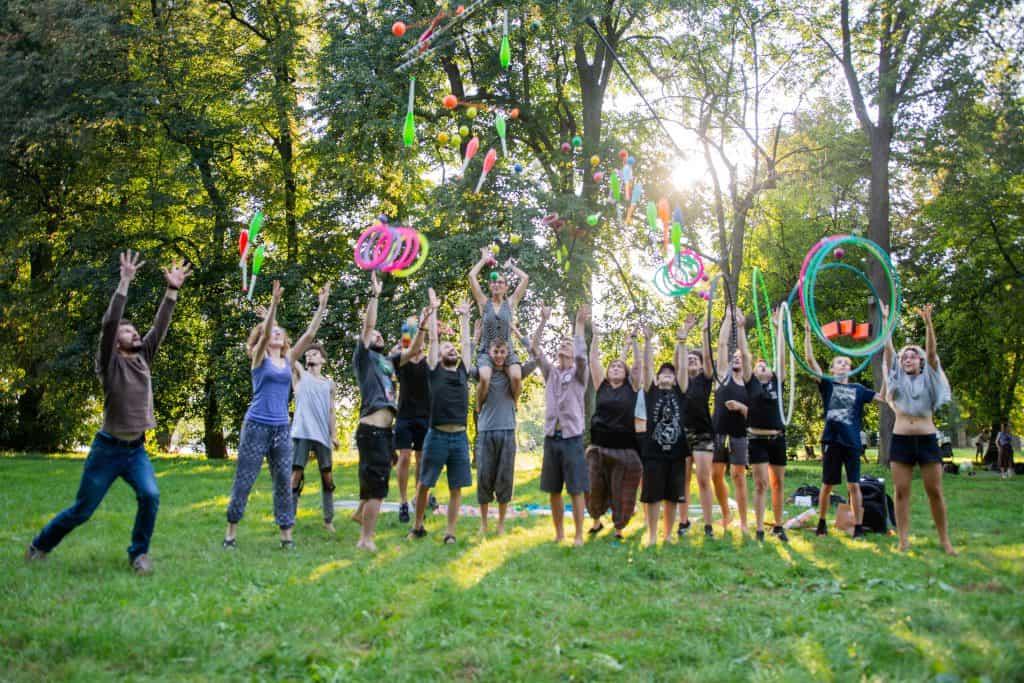 Siedemnaście osób rzuca w górę cyrkowe rekwizyty: maczugi i piłki do żonglowania, obręcze żonglerskie, hula hop. W tle drzewa