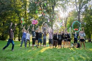 Siedemnaście osób rzuca w górę cyrkowe rekwizyty: maczugi i piłki do żonglowania, obręcze żonglerskie, hula hop. W tle drzewa