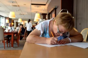 kobieta pochyla się nad stołem. W dłoni ma długopis. Trzyma go na kartce W tle ludzie siedzący przy stołach.