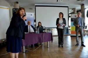 kobieta trzyma mikrofon w dłoni. W tle inni ludzie. W tle ekran multimedialny.