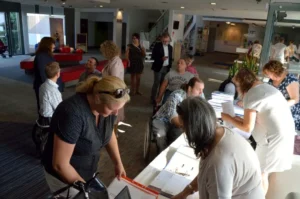 Ludzie pochylają sie nad stołami. Są w pomieszczeniu. Na stołach różne papiery.