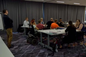 Ludzie siedzą przy stołach. Są w pomieszczeniu. Przed nimi na środku stoi mężczyzna. Ma założone ręce na siebie,