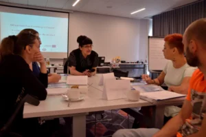 Ludzie siedzą przy stole. Nachylają sie ku sobie. W tle prezentacja multimedialna.