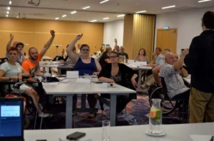 Ludzie siedza przy stołach. Podnoszą swoje ręce ku górze. Są uśmiechnieci.