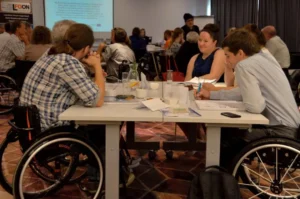 Ludzie siedza przy stołach. Są osoby na wózkach inwalidzkich. Twarze ludzi są zwrócone ku sobie