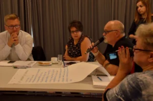 Ludzie siedza przy stołach. Męzczyzna trzyma mikrofon w dłoni. Przed nim duża kartka na stole.