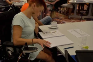 Kobieta na wózku siedzi przy stole. Pochyla się nad broszura.