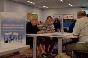 Ludzie siedzą przy stole. Twarzami są skierowani ku sobie W tle baner reklamowe.