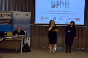 Kobiety stoją na scenie. Trzymaja mikrofony w dłoniach. W tle prezentacja na ekranie multimedialnym jest wyświetlana.