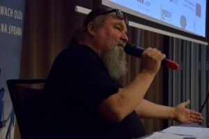 Mężczyzna siedzi przy stole. W dłoni trzyma mikrofon. Druga ręka jest uniesiona.