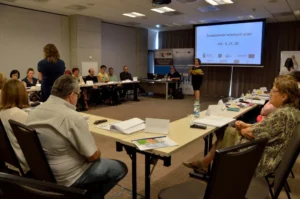 Ludzie siedzą przy stołach. Na środku sali stoją kobiety. W tle ekran multimedialny.