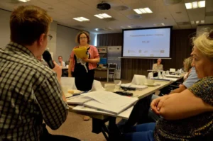 Ludzie siedzą prz stole. W tle prezentacja na ekranie multimedialnym