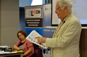 Mężczyzna stoi. W dłoni trzyma książkę. Palcem wskazującym wskazuje na kartkę.