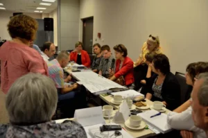 Ludzie siedzą przy stole. Ich twarze są zwrócone ku sobie.